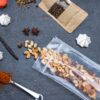 Bild zeigt einen durchsichtigen Doypack®-Beutel, gefüllt mit Nüssen und Trockenfrüchten, umgeben von Gewürzen, Baisers und anderen Lebensmitteln. Der Beutel ist luftdicht und aus PE/VOH-PE mit einer EVOH-Schicht hergestellt, die Schutz vor Sauerstoff und Feuchtigkeit bietet. Der Beutel ist ideal für verschiedene Food- und Non-Food-Zwecke, wiederverschließbar und recycelbar.